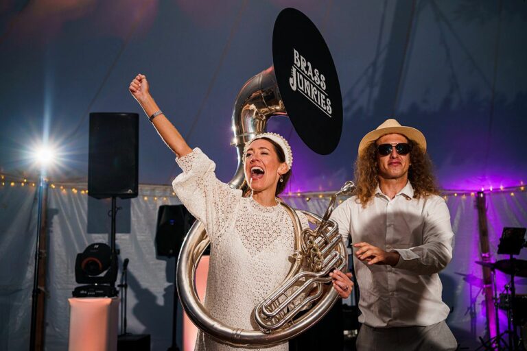 A joyous woman in a white dress plays a sousaphone decorated with a "Brass Junkies" sign. She smiles widely and raises her left arm in celebration. Beside her, a man in a white shirt and hat, wearing sunglasses, dances energetically. Festive lighting sets the scene. Image by Documentary Wedding Photographer Lewis Bishop Photography.