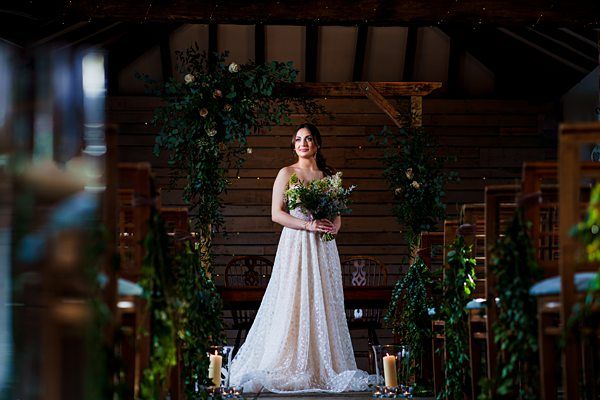A bride stands in the center aisle of a rustic, dimly lit venue, holding a bouquet of flowers. She is wearing a white, lace wedding gown. The aisle is adorned with greenery and candles, and there's a floral arch behind her. The wooden interior creates a cozy atmosphere. Image by Documentary Wedding Photographer Lewis Bishop Photography.