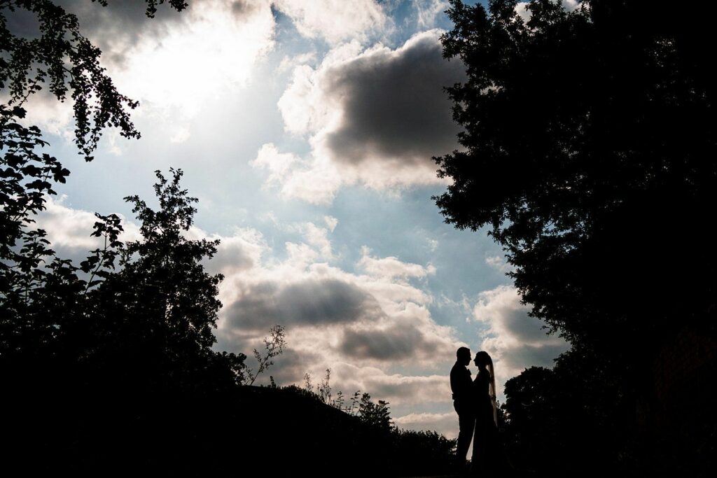 A silhouette of a couple standing closely together against a backdrop of a partly cloudy sky. Tall trees frame either side of the image, and the sunlight filters through the clouds, casting a soft glow on the scene. Image by Documentary Wedding Photographer Lewis Bishop Photography.