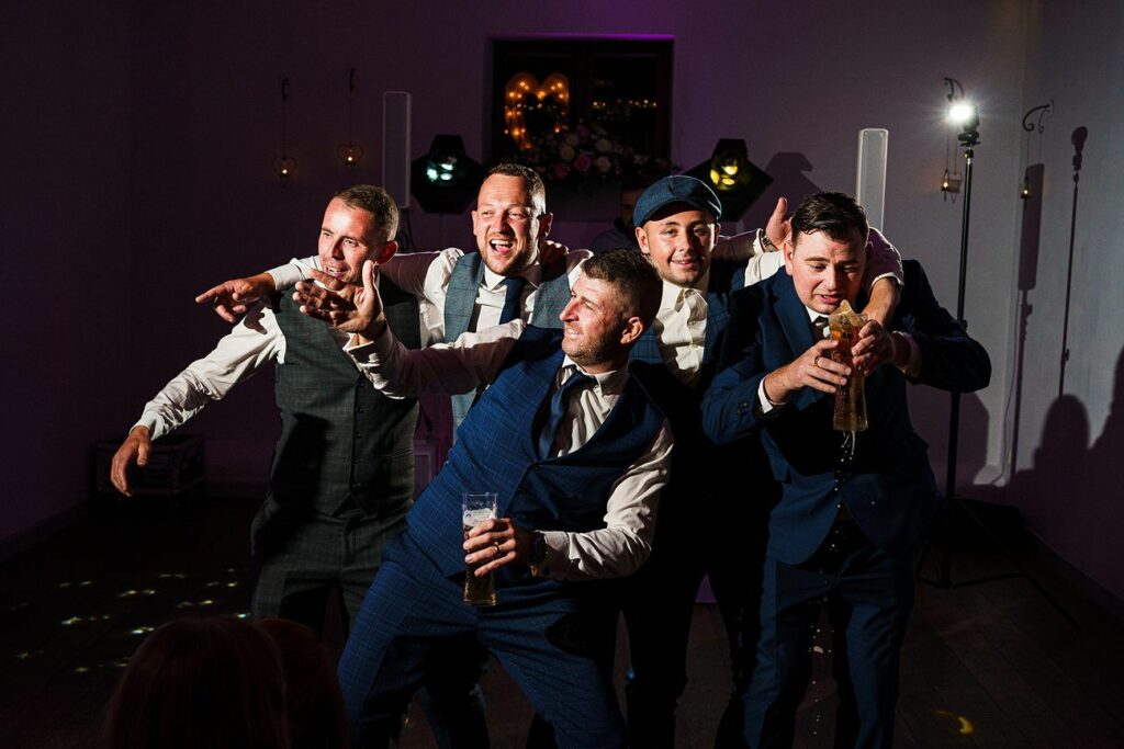 A group of five men in suits are joyfully dancing and laughing. Some hold drinks, and all appear to be having a great time. They are illuminated by party lights in an otherwise dimly-lit room, giving the scene a lively and celebratory atmosphere. Image by Documentary Wedding Photographer Lewis Bishop Photography.