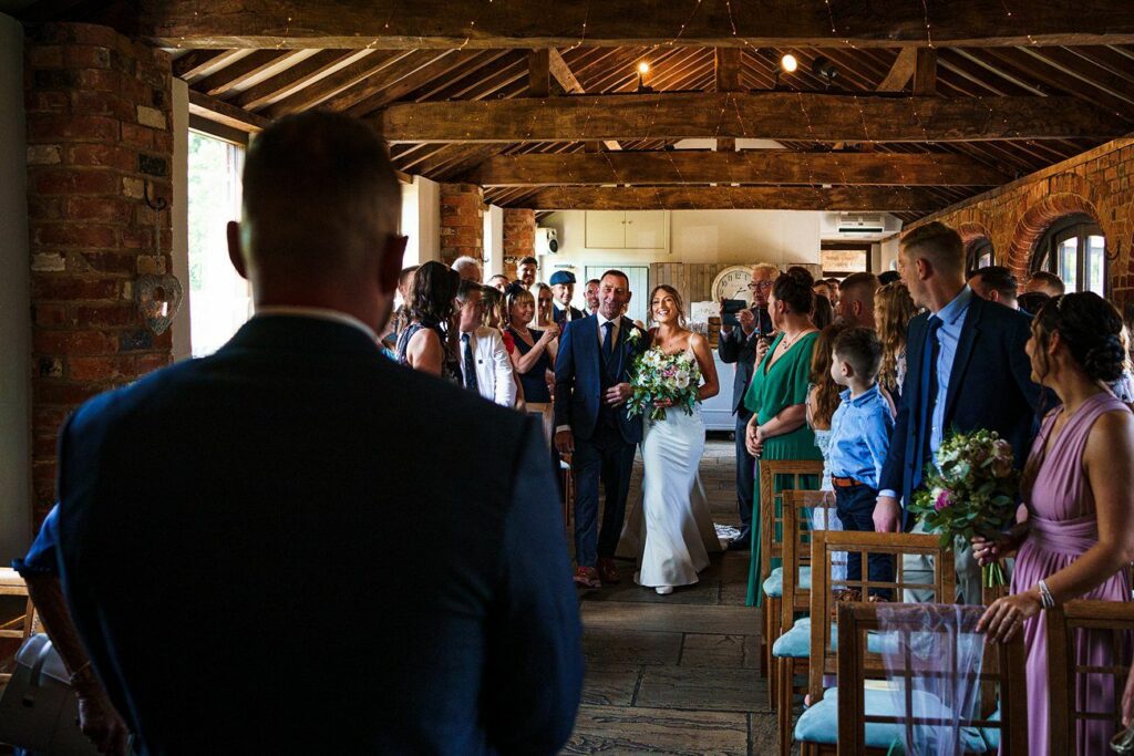A bride, dressed in a white gown and holding a bouquet, walks down the aisle with her father in a rustic indoor venue. Guests are standing and watching them, with some seated. The room has wooden beams, exposed brick walls, and soft lighting. Image by Documentary Wedding Photographer Lewis Bishop Photography.