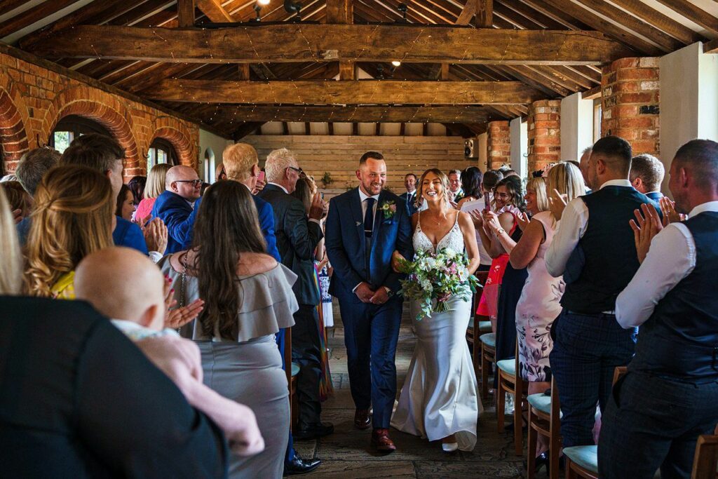 A newly married couple, dressed in a blue suit and a white wedding gown, walks down the aisle together in a rustic venue filled with wooden beams and brick walls. Guests on both sides, including a mother holding a baby, clapping and smiling in celebration. Image by Documentary Wedding Photographer Lewis Bishop Photography.