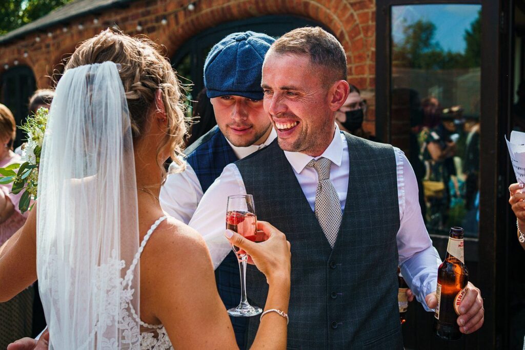 A bride in a white dress and veil stands facing two men in vests and ties. One man wears a blue flat cap, and the other smiles broadly while holding a beer. The bride holds a glass of rosé wine. They are outside a brick building, celebrating. Image by Documentary Wedding Photographer Lewis Bishop Photography.