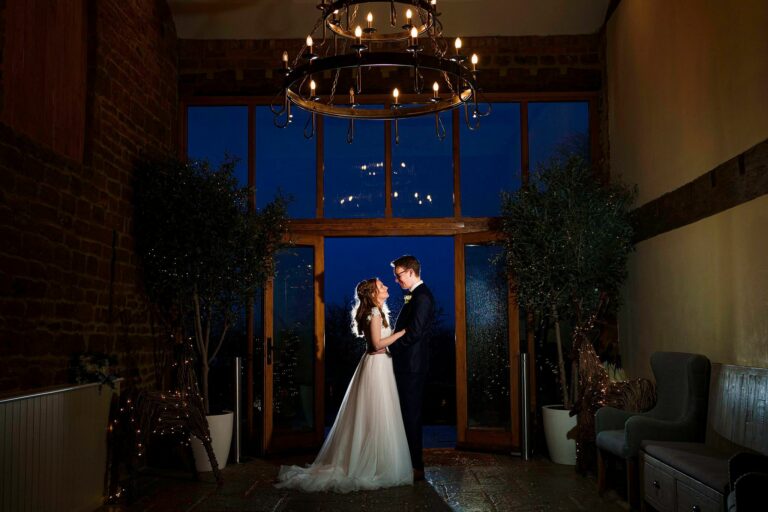 A bride and groom share a moment under a dimly lit, elegant chandelier in an intimate, rustic room with exposed brick walls. The large glass doors behind them reveal a deep, dark blue night sky. The bride is in a white dress, and the groom is in a dark suit. Image by Documentary Wedding Photographer Lewis Bishop Photography.