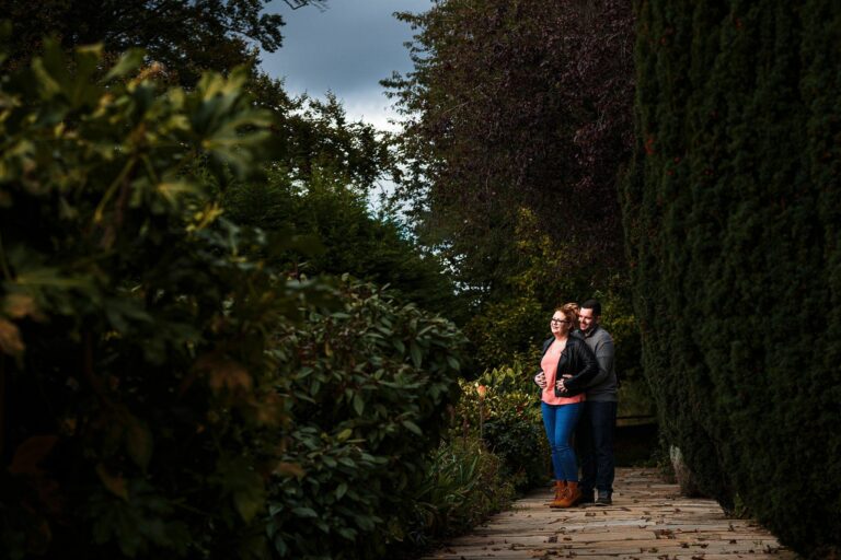 A couple stands close together, with the man wearing a leather jacket and the woman wearing a pink sweater, denim, and glasses. They are on a narrow stone path surrounded by dense greenery and tall trees, under a partly cloudy sky. Image by Documentary Wedding Photographer Lewis Bishop Photography.