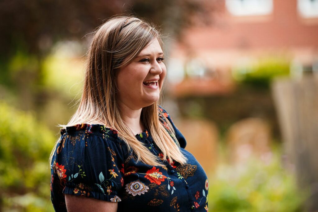 A woman with long light brown hair, wearing a dark floral blouse, is smiling. She is outdoors with blurred greenery and a building in the background. She is captured in a candid, side profile view, exuding a cheerful demeanor. Image by Documentary Wedding Photographer Lewis Bishop Photography.