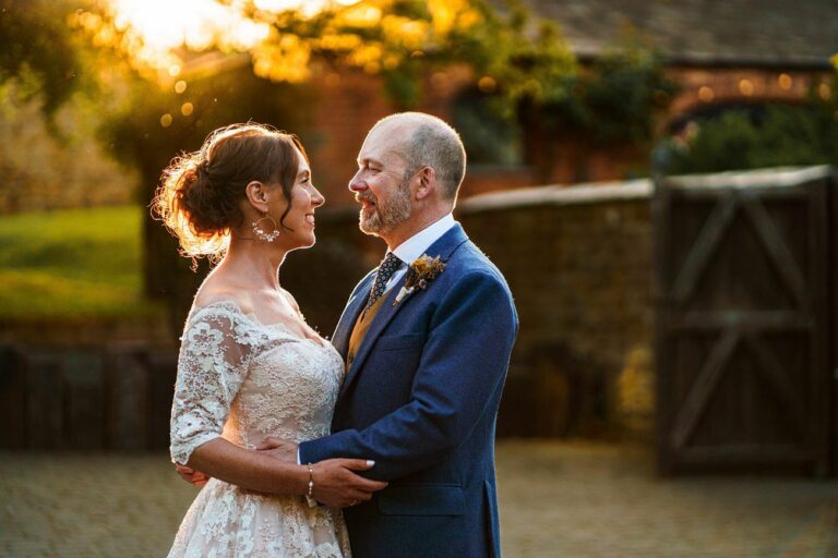A couple in wedding attire stand facing each other and smiling, illuminated by warm, golden sunlight. They are outdoors near a stone building and large wooden gates in the background. Image by Documentary Wedding Photographer Lewis Bishop Photography.