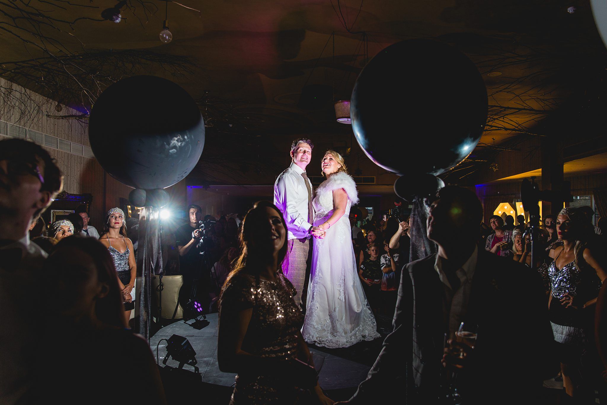 A couple is standing on a stage, holding hands, and smiling while wearing formal wedding attire. They are surrounded by an audience, with two large black balloons framing them in the foreground. The scene is dimly lit, creating a spotlight effect on the couple. Image by Documentary Wedding Photographer Lewis Bishop Photography.