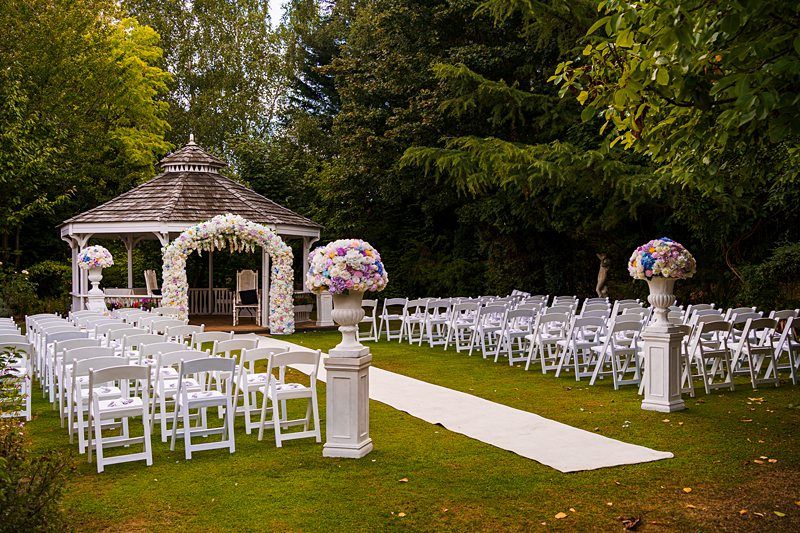 An outdoor wedding venue with a white gazebo adorned with floral decorations. White folding chairs are arranged in rows on either side of an aisle covered with a white runner. Two large floral arrangements stand at the entrance to the aisle. Trees surround the scene. Image by Documentary Wedding Photographer Lewis Bishop Photography.