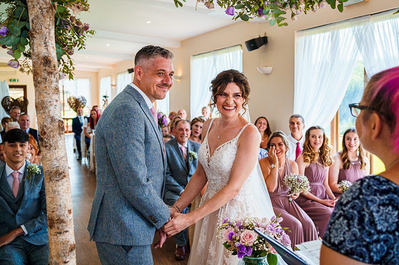 A smiling bride and groom hold hands during their wedding ceremony in a bright, flower-adorned room filled with seated guests. The bride wears a white lace dress, and the groom wears a light gray suit. Bridesmaids in pink dresses sit joyfully behind the couple. Image by Documentary Wedding Photographer Lewis Bishop Photography.
