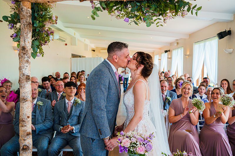 A couple shares a kiss at the altar during their wedding ceremony. The bride is wearing a white dress and holding a bouquet of flowers, while the groom is in a grey suit. Guests, dressed in formal attire, are seated behind them, smiling and clapping. Image by Documentary Wedding Photographer Lewis Bishop Photography.