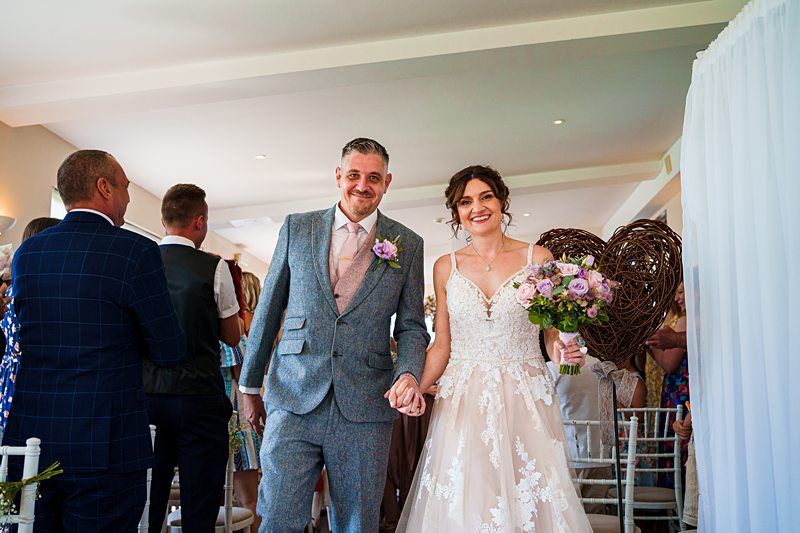 A bride in a white lace dress and a groom in a gray suit hold hands and smile as they walk down the aisle. The bride holds a bouquet of flowers. Guests in formal attire stand and smile in the background. The setting is a well-lit indoor space. Image by Documentary Wedding Photographer Lewis Bishop Photography.