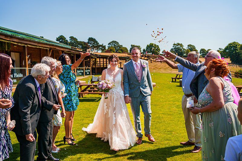 A bride and groom walk hand in hand outside while being showered with flower petals by a group of guests. The bride wears a white gown and holds a bouquet, while the groom wears a light blue suit with a pink tie. The event appears to be a wedding celebration. Image by Documentary Wedding Photographer Lewis Bishop Photography.