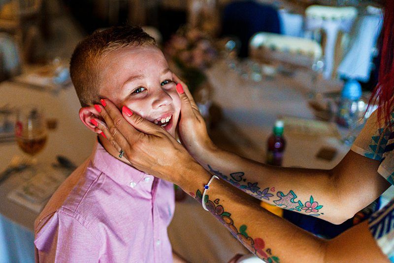 A smiling young boy in a pink shirt is having his cheeks gently squeezed by an adult with bright red nails and colorful tattoos on their arm. The background shows a blurred view of a dining table set for a meal. Image by Documentary Wedding Photographer Lewis Bishop Photography.