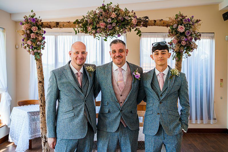 Three men in gray suits stand smiling under a wooden wedding arch adorned with flowers. The man in the center wears a pink vest and tie, while the other two wear matching ties and boutonnieres. The room is well-lit, with sunlight flooding in through sheer curtains. Image by Documentary Wedding Photographer Lewis Bishop Photography.