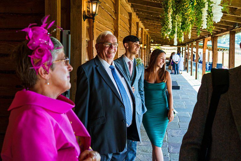 A group of people at an outdoor event under a wooden pergola. A woman in a pink dress and fascinator, an older man in a suit and tie, a man in a cap and blazer, and a woman in a teal dress are smiling and talking. Hanging flowers adorn the pergola. Image by Documentary Wedding Photographer Lewis Bishop Photography.