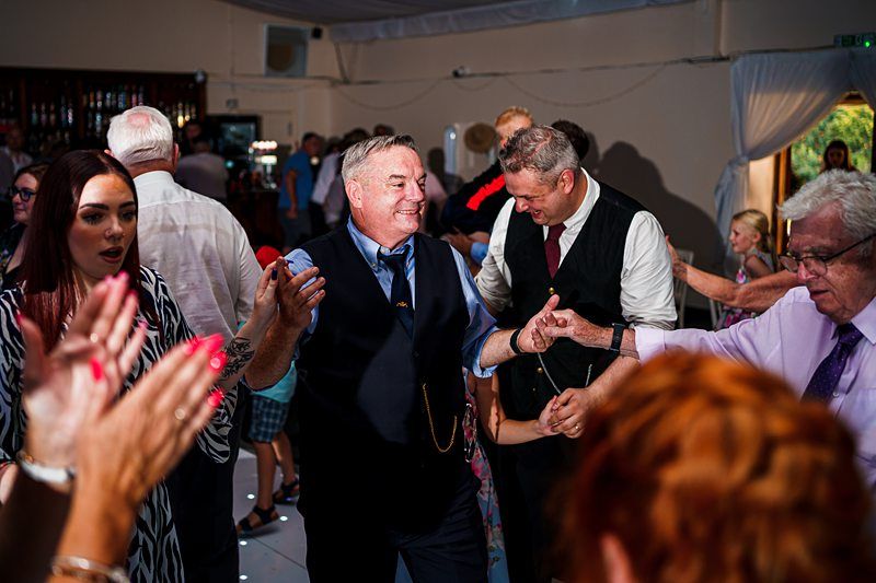 A group of people dance joyfully in a brightly lit indoor setting. Two men in vests and ties, one with grey hair, hold hands with others while dancing. A woman with auburn hair and a striped dress claps along. The background shows others participating and mingling. Image by Documentary Wedding Photographer Lewis Bishop Photography.