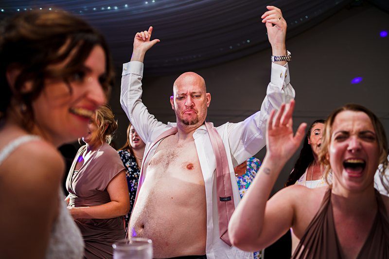 A group of people dancing joyfully at an event. A bald man in an unbuttoned shirt raises his arms with a playful expression, while two women laugh and smile. The vibrant atmosphere is accentuated by dim, colorful lighting. Image by Documentary Wedding Photographer Lewis Bishop Photography.