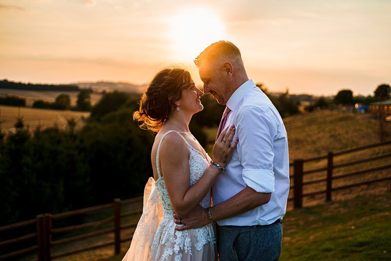 A couple embraces and gazes into each other's eyes during sunset. The woman wears a white, lace wedding dress, and the man wears a white shirt with rolled-up sleeves and a tie. The sun sets behind them, casting a warm glow over the rural landscape. Image by Documentary Wedding Photographer Lewis Bishop Photography.