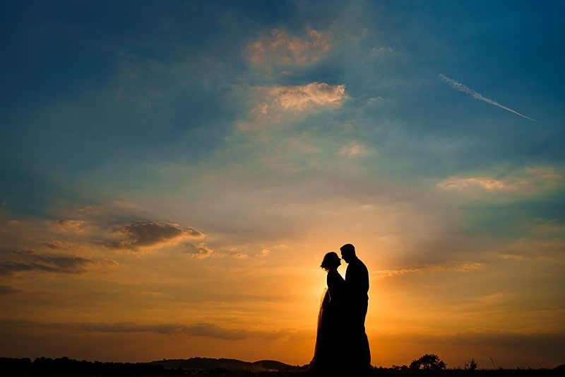 A silhouette of a couple standing close, facing each other against a vibrant sunset sky with hues of orange and blue. The sun is near the horizon, creating a warm glow around them. Clouds and a distant tree line are visible in the background. Image by Documentary Wedding Photographer Lewis Bishop Photography.