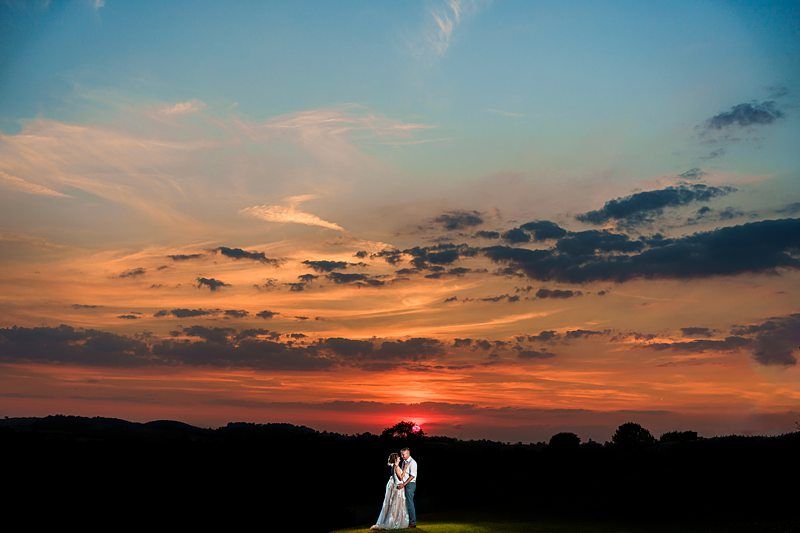 A bride and groom stand close together in a field as the sun sets behind them. The sky is filled with vibrant colors ranging from blue to orange, with wispy clouds adding texture. The couple is illuminated, contrasting against the darkening landscape. Image by Documentary Wedding Photographer Lewis Bishop Photography.