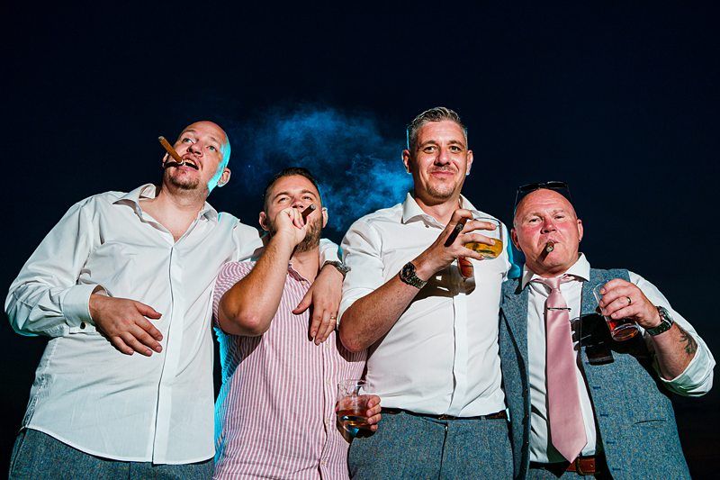 Four men stand together against a dark blue sky, each holding a cigar and drinks. They are dressed in casual formal attire, with open collars and rolled sleeves, appearing relaxed and cheerful. Smoke from the cigars is visible in the background. Image by Documentary Wedding Photographer Lewis Bishop Photography.