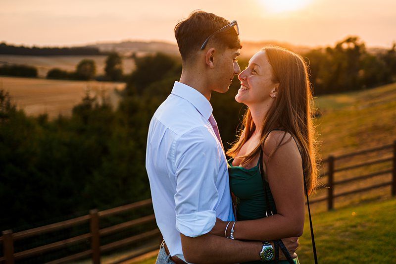 A couple stands in a close embrace, smiling at each other as the sun sets behind them. They are outdoors, with a scenic view of fields, trees, and distant hills. The atmosphere is warm and romantic, with a soft, golden light. Image by Documentary Wedding Photographer Lewis Bishop Photography.
