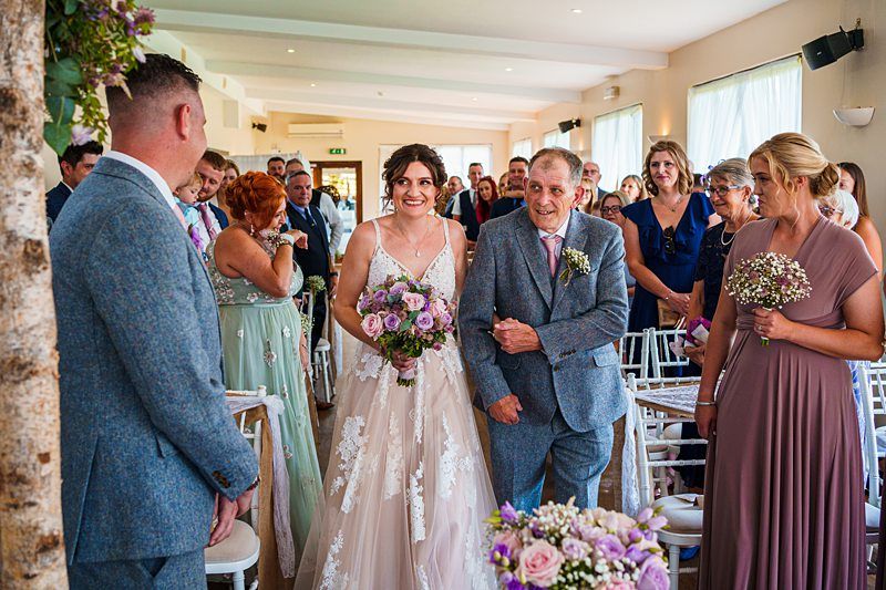 A bride in a white gown, carrying a bouquet, walks down the aisle accompanied by an older man in a grey suit, presumably her father. The aisle is lined with guests, and at the end, a man waits in a light blue suit. The setting appears to be an indoor wedding venue. Image by Documentary Wedding Photographer Lewis Bishop Photography.