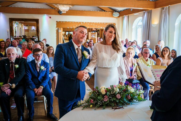 A couple stands at an altar holding hands during their wedding ceremony. The groom is wearing a blue suit and the bride is in a white dress. Guests are seated behind them, watching the ceremony. A floral arrangement decorates the table in front of them. Image by Documentary Wedding Photographer Lewis Bishop Photography.