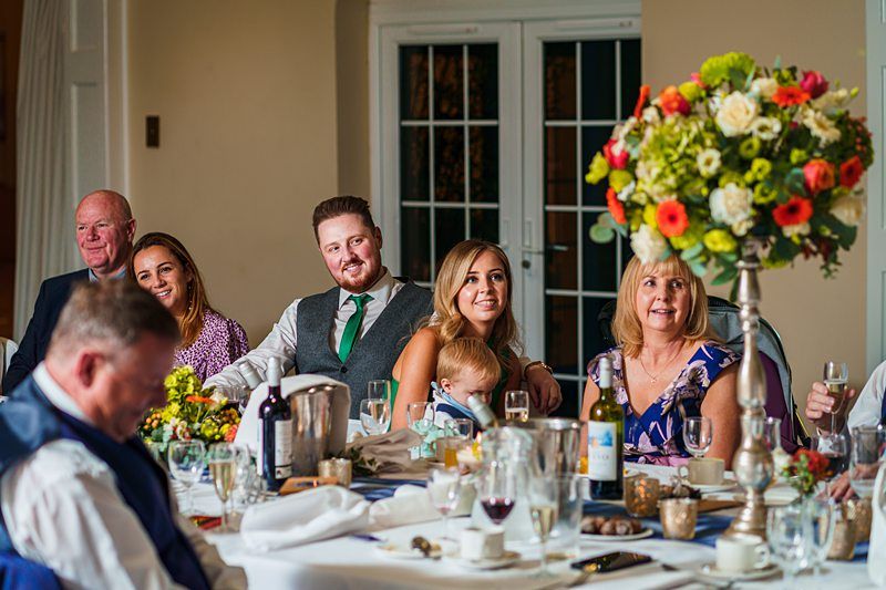 A group of people, including men, women, and a child, are seated at a table set for a meal, smiling at the camera. The table is decorated with glasses, dishes, a bottle of wine, and a tall floral centerpiece with colorful flowers. A glass door is visible in the background. Image by Documentary Wedding Photographer Lewis Bishop Photography.