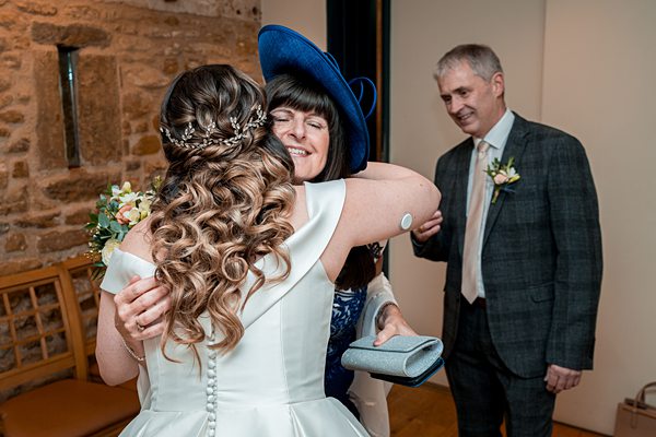 Bride hugging guest at wedding, smiling man watching.
