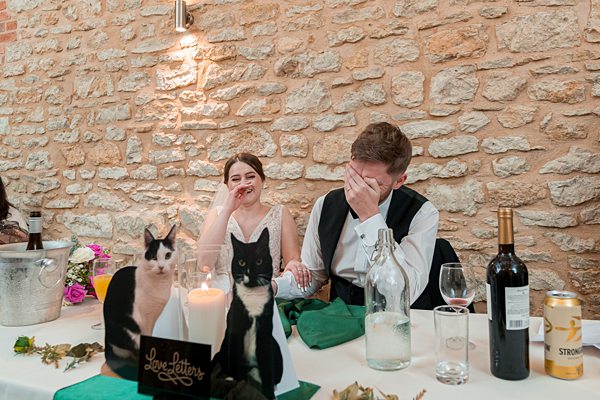 Wedding couple laughing near cats at reception table.
