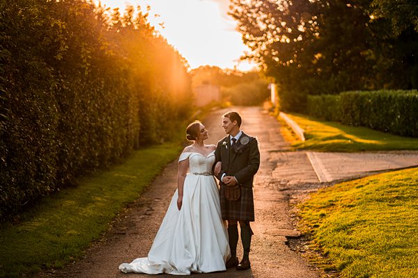 Couple in wedding attire at sunset on tree-lined path.