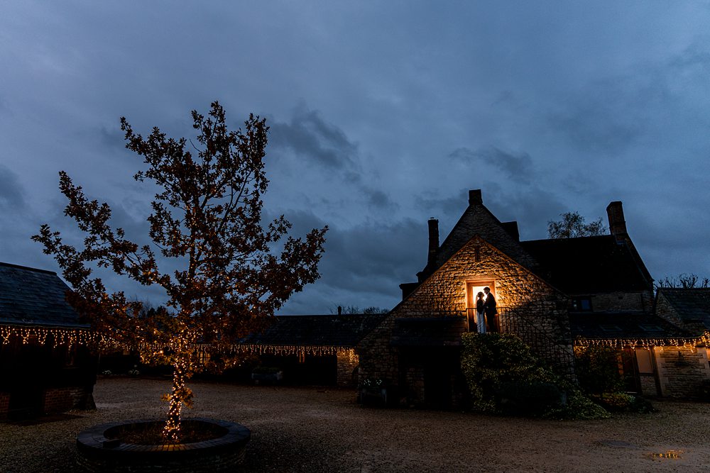Twilight at illuminated cottage with festive lights and tree.