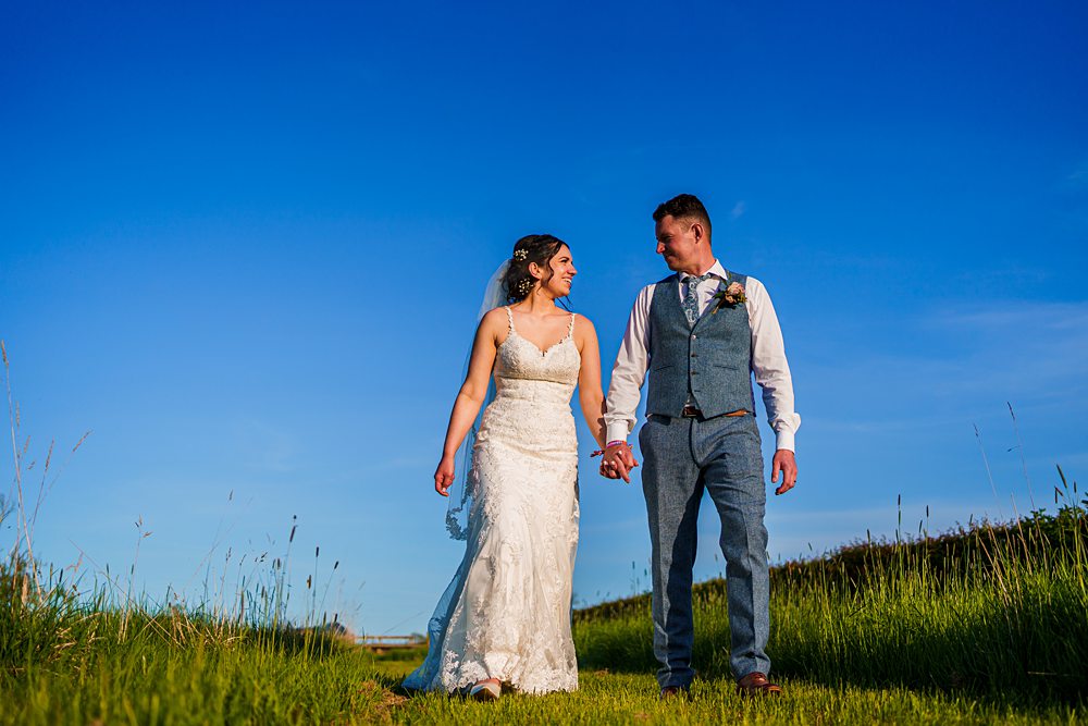 Couple in wedding attire walking through a sunny meadow.