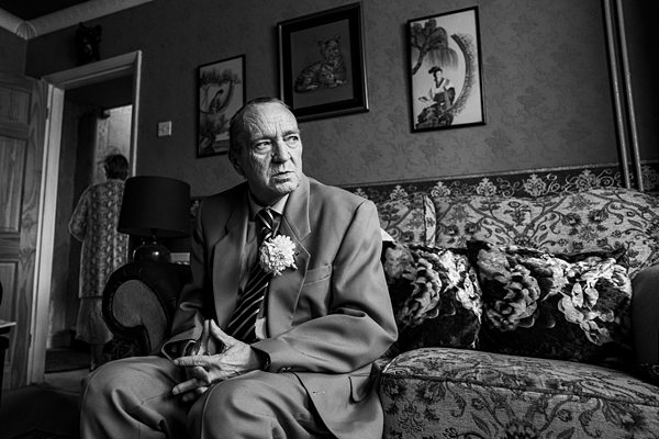 Elderly man in suit sitting on patterned sofa, contemplative.