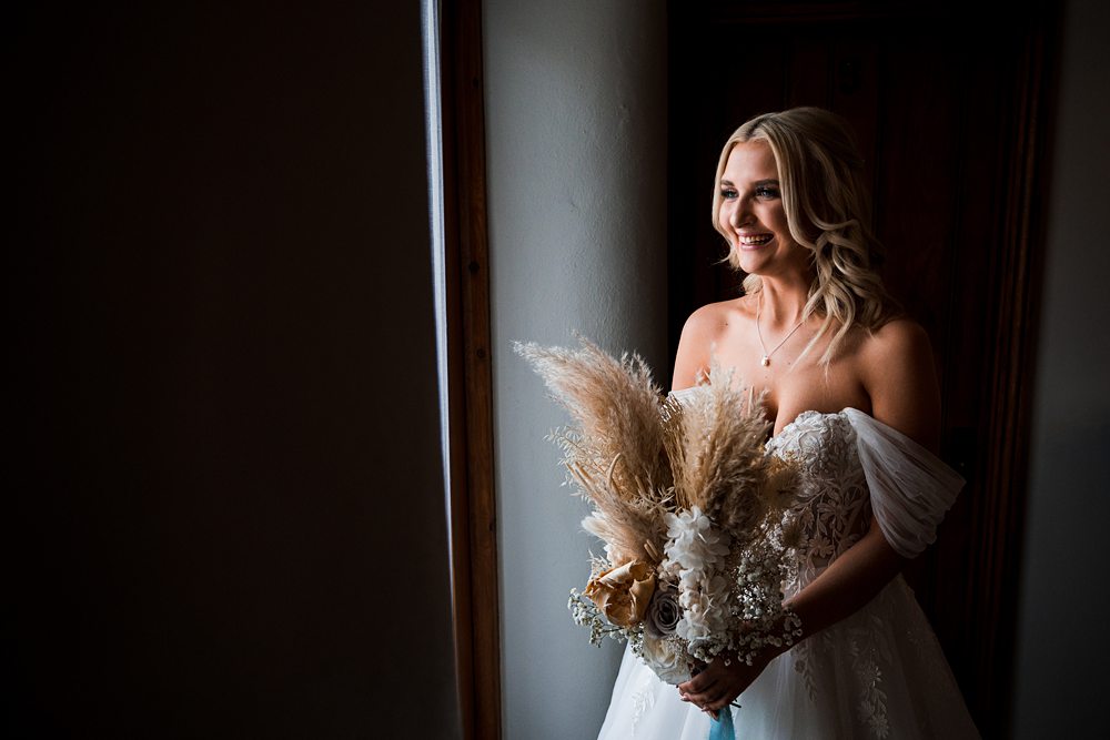 Bride holding bouquet, smiling, standing by window.