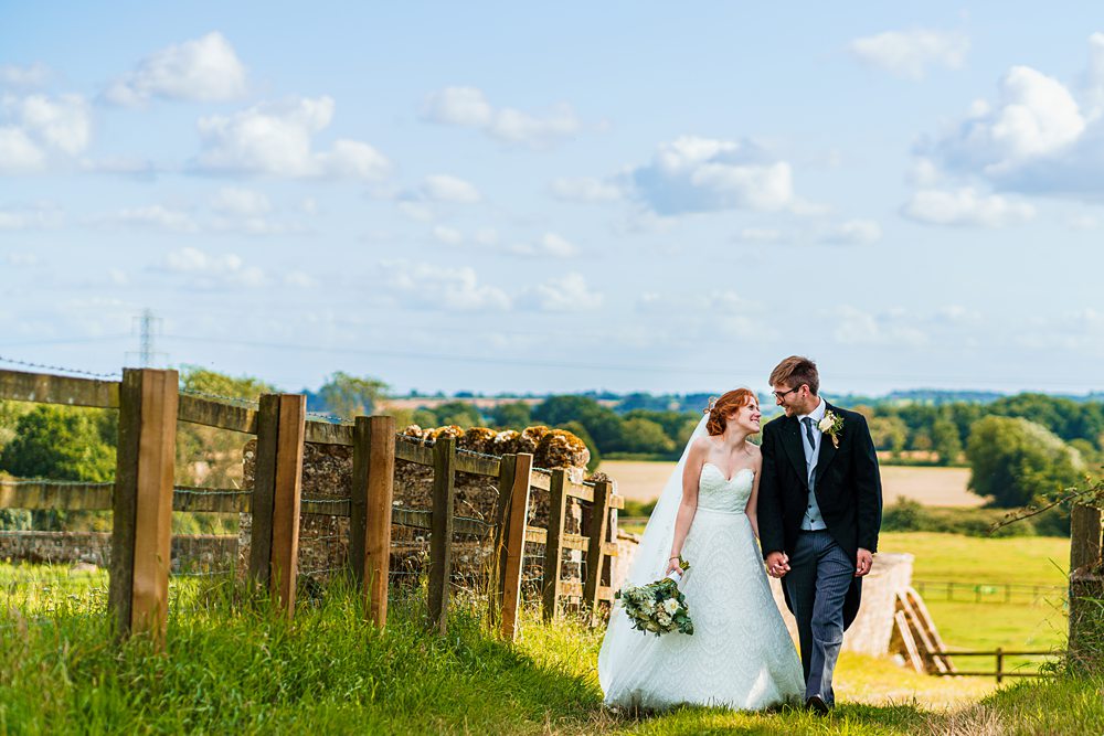 Bride and groom walking by countryside fence.