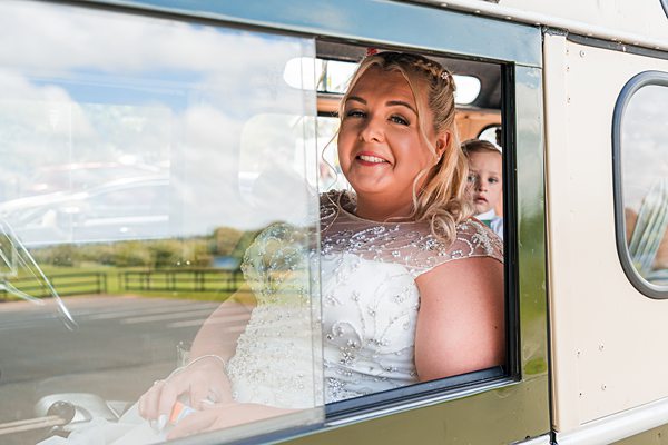 Bride smiling in car with child peeking in background.