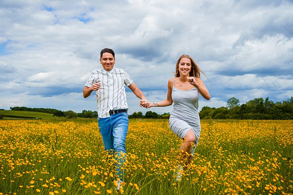 Couple running through wildflower meadow under cloudy sky.