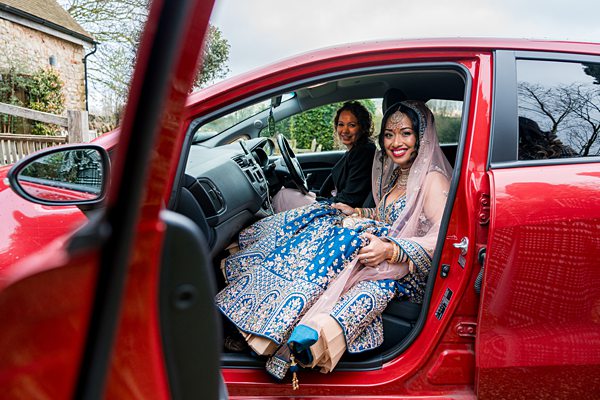 Bride in traditional attire inside a red car smiling.