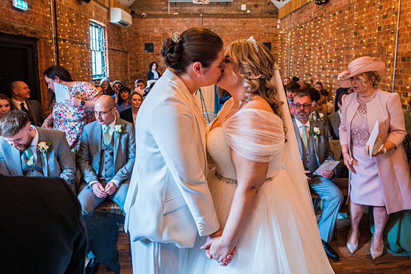 Brides kissing at wedding ceremony with guests watching.