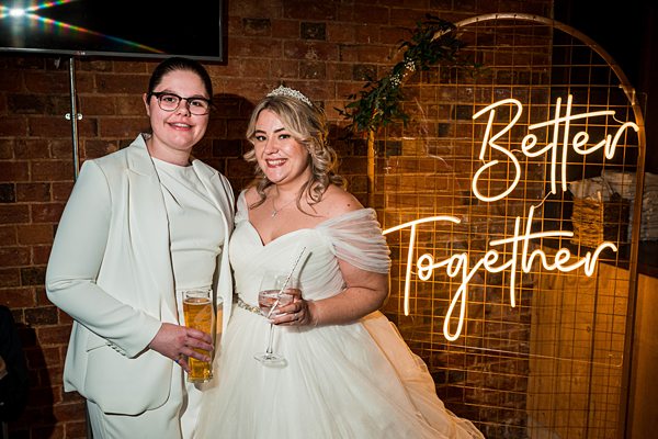 Two brides celebrating wedding with "Better Together" neon sign.