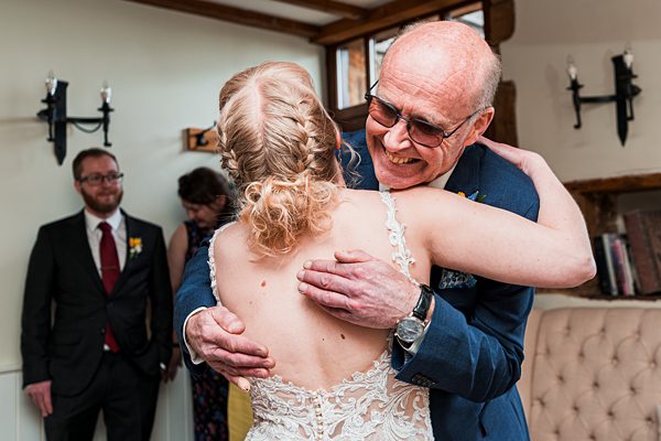 A man with glasses, wearing a blue suit, embraces a woman in a white lace dress with a braided hairstyle, both smiling. Two individuals, a man in a black suit and a woman in a floral dress, stand in the background. The setting appears to be indoors. Image by Documentary Wedding Photographer Lewis Bishop Photography.