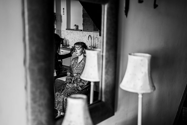 A woman wearing a floral robe sits with her eyes closed, reflected in a wall mirror. The black and white image showcases a cozy room with dim lighting, a table lamp, and a tiled backsplash behind her. The ambiance is serene. Image by Documentary Wedding Photographer Lewis Bishop Photography.