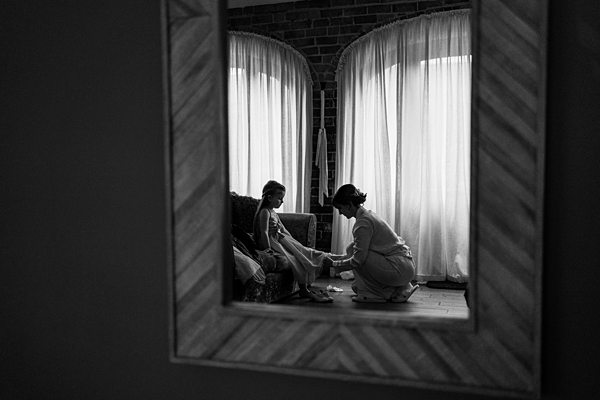 A black-and-white photo shows a woman kneeling and helping a young girl with her shoes. They are in a room with large, curtained windows that let in soft light. The image is captured through a mirror on a wall, giving it a framed perspective. Image by Documentary Wedding Photographer Lewis Bishop Photography.