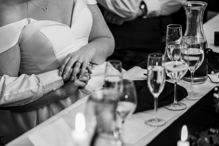 A black and white photo shows a close-up of a couple holding hands at a table set for a meal. The bride, in a white dress, is sitting beside the groom. Several glasses of wine and water, as well as a candle, are visible on the table. Image by Documentary Wedding Photographer Lewis Bishop Photography.