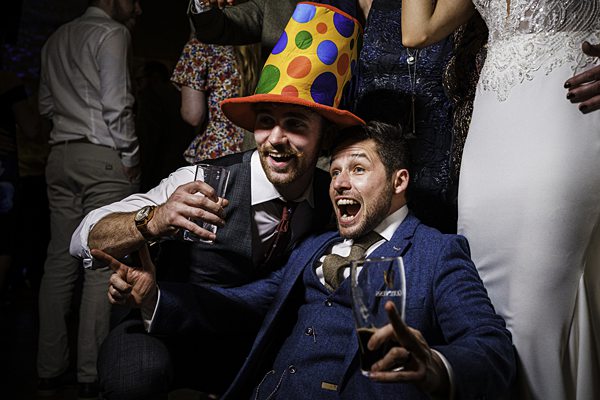 Two men at a lively event, both smiling and holding drinks. The man on the left is wearing a colorful, polka-dotted hat, while the man on the right is exclaiming with excitement. They are dressed in suits and surrounded by other partially visible attendees. Image by Documentary Wedding Photographer Lewis Bishop Photography.