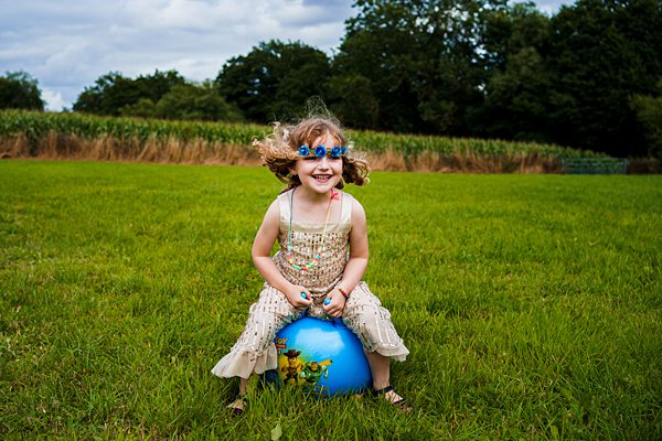 A young girl with curly hair, wearing a floral headband and a light-colored dress, is joyfully bouncing on a large blue ball in a grassy field. The sky is partly cloudy, and trees can be seen in the background. Image by Documentary Wedding Photographer Lewis Bishop Photography.