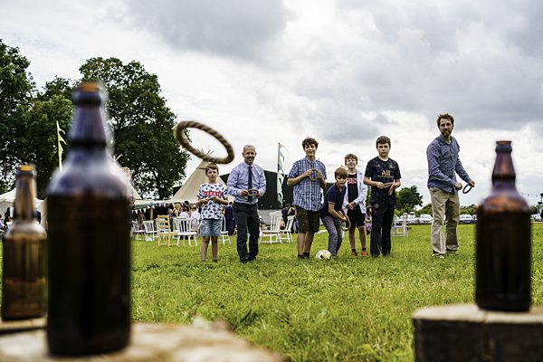 A group of seven people, including children and adults, play ring toss on a grassy field. Bottles are arranged in the foreground as targets. A large tent and chairs are visible in the background under a cloudy sky. Everyone appears engaged and focused. Image by Documentary Wedding Photographer Lewis Bishop Photography.